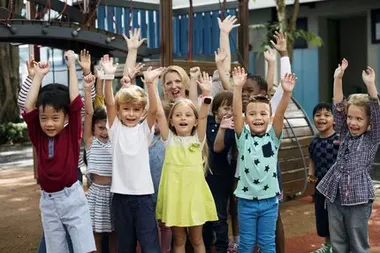 Jubelnde Kinder, die die Arme in die Luft strecken und lachen. Im Hintergrund ein Kinderspielplatz.