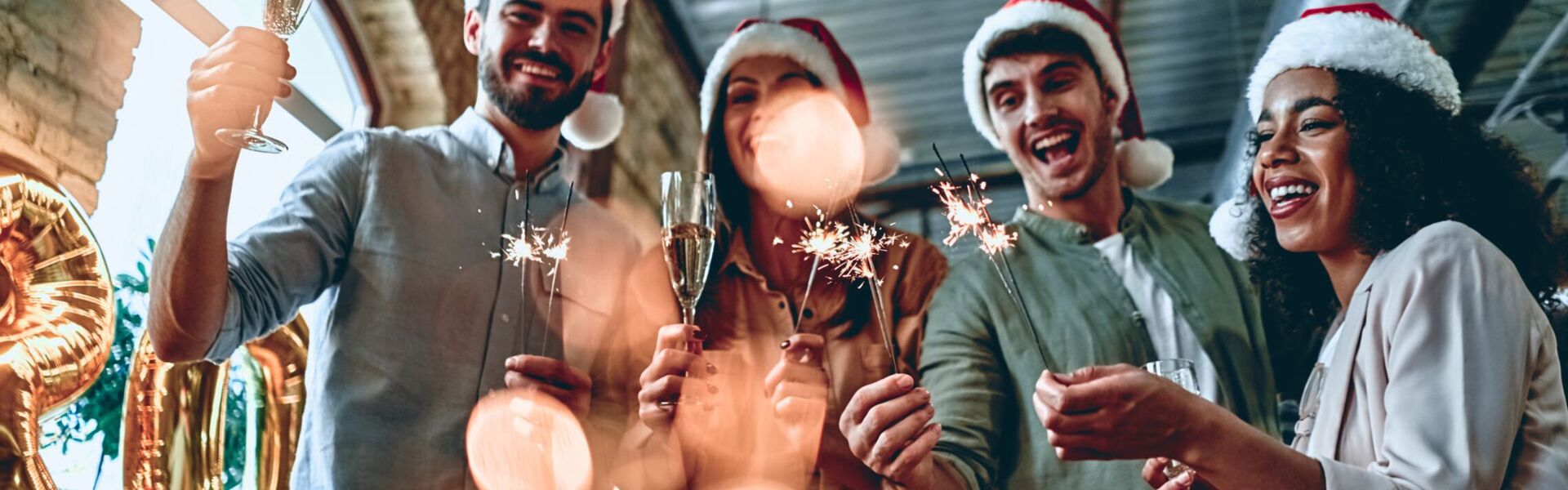 Vier Personen mit Weihnachtsmützen, Wunderkerzen, und Sektgläsern am feiern.